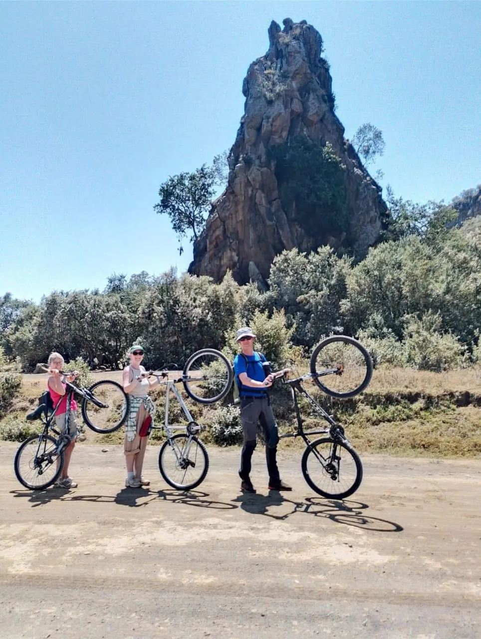 Three travelers lifting bicycles near a tall rock formation